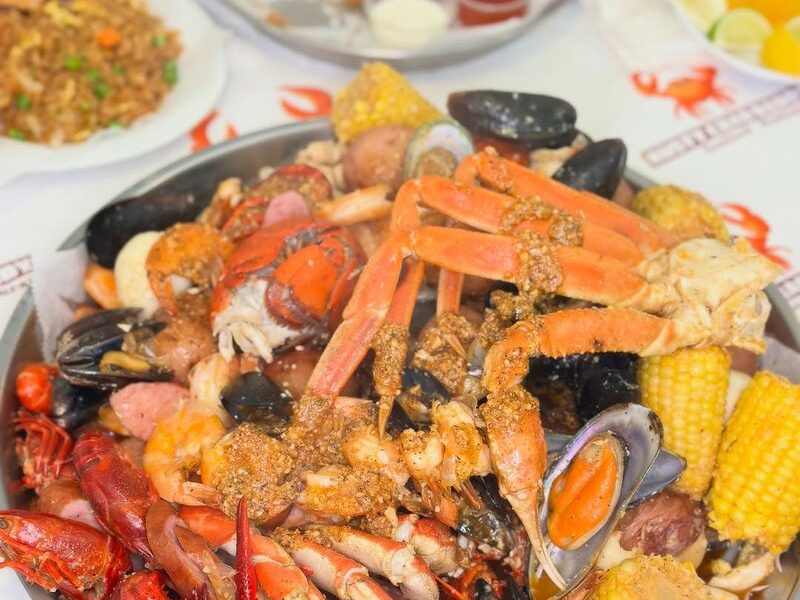A seafood boil tray with crab legs, crawfish and corn, with fried chicken, rice and drinks behind