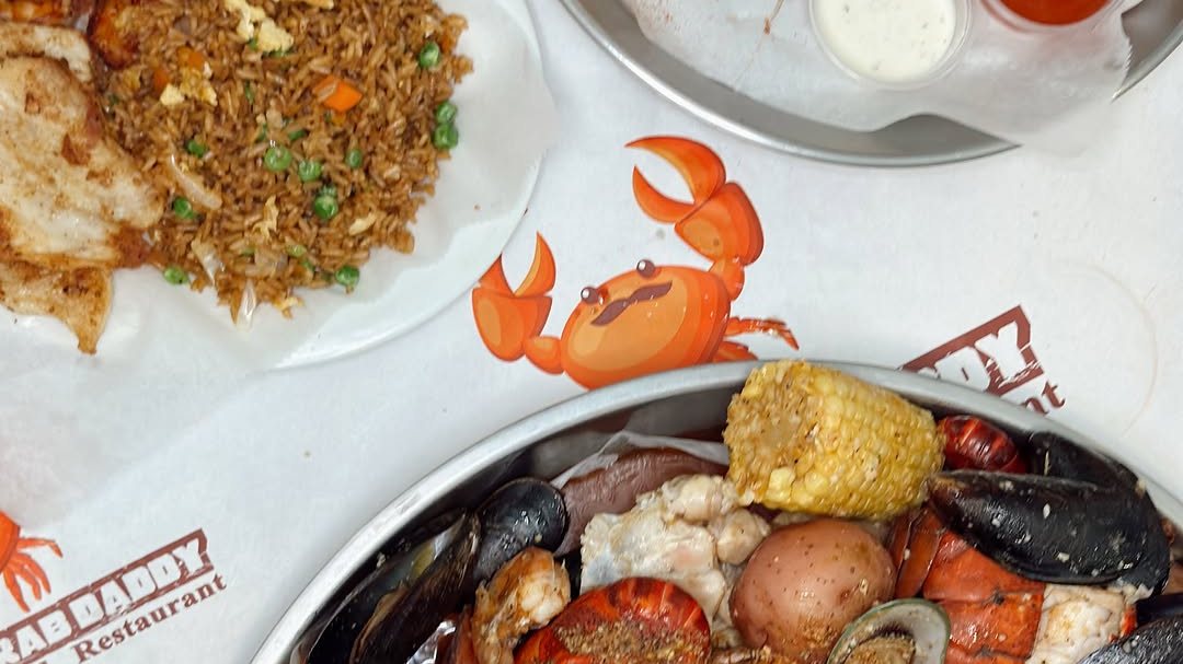 A seafood boil tray with mussels, corn and potato beside a plate of fried rice on Rusty Crab Daddy's crab-print table paper